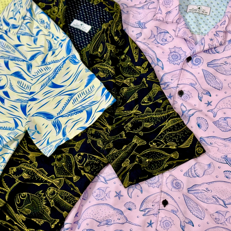 Three ocean-themed shirts from Tropical Seas Clothing with marine life designs. The pink shirt shows dolphins and seashells, while the darker ones feature contrasting marine patterns. Perfect for an eco-friendly coastal style.