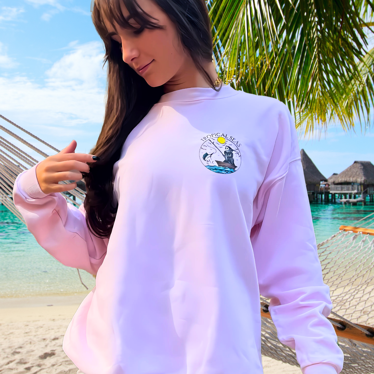 “Ashtin Earle wearing a pastel pink Tropical Seas Clothing sweatshirt with an ocean-themed graphic, standing by a tropical beach with palm trees and overwater bungalows in the background. Showcasing sustainable beachwear that promotes ocean conservation.