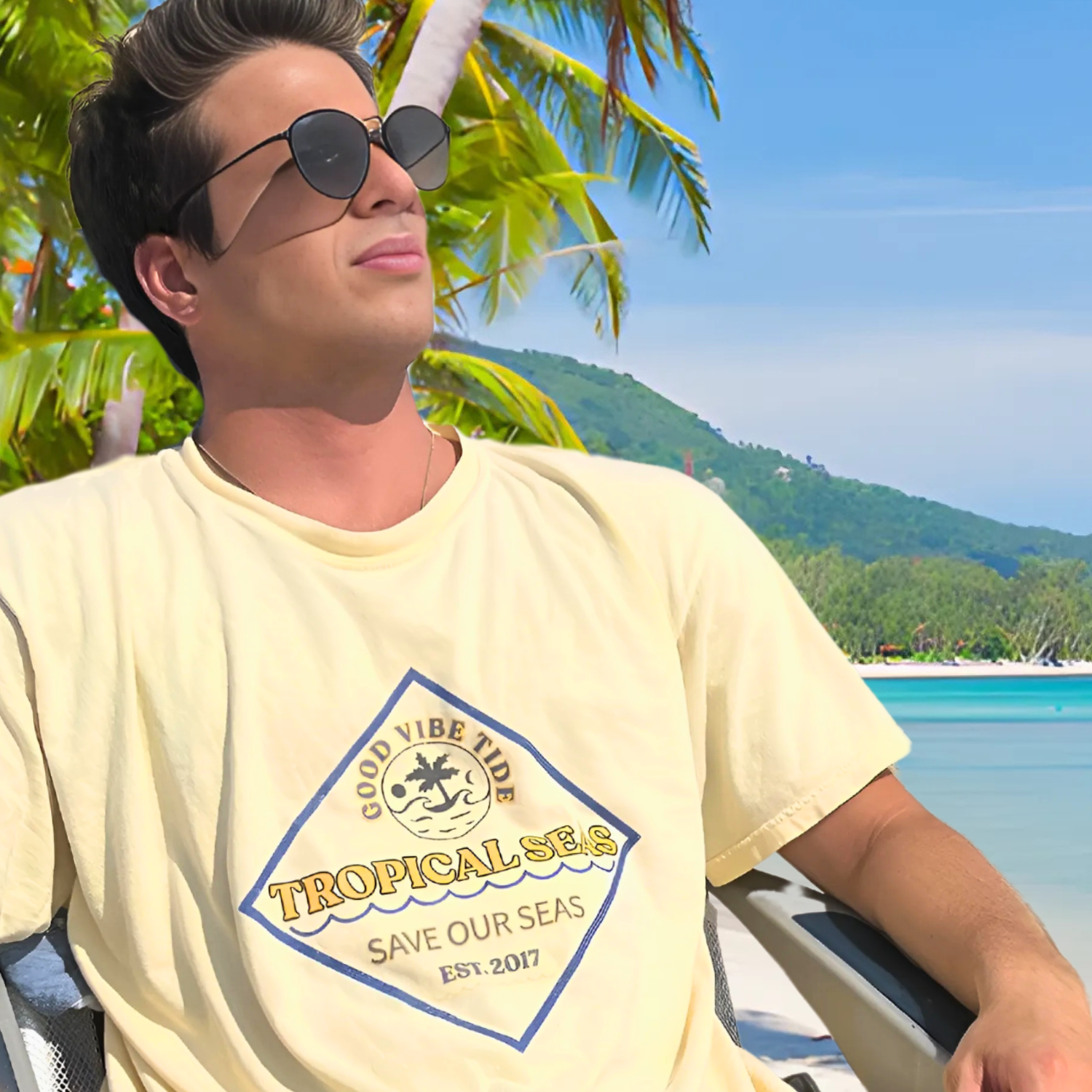 Man wearing Tropical Seas Clothing Men’s T-Shirt with Good Vibe Tide design, relaxing by a tropical beach, embodying sustainable fashion and ocean conservation.